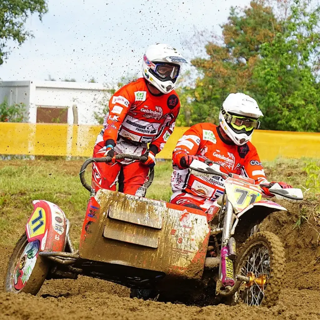 Das Motocross-Gespann-Team Marcus Prokesch & Nick Kutschke (MSC Geisleden) in orange-weißen Rennanzügen und Helmen fährt ihr stark verschlammtes Fahrzeug mit der Startnummer 71 auf einer schlammigen Strecke, wobei Erde aufgewirbelt wird.