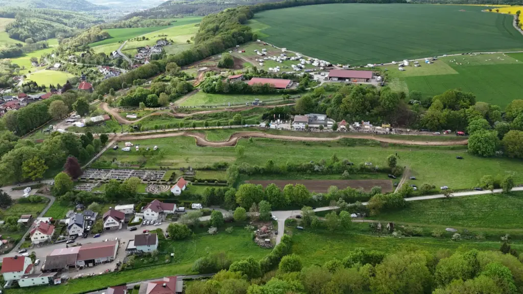Luftaufnahme der Motocross-Strecke des MSC Geisleden, eingebettet in eine ländliche Landschaft mit grünen Feldern, Wäldern und einem angrenzenden Dorf. Gebäude, Fahrzeuge und Menschen säumen die kurvenreiche Strecke.