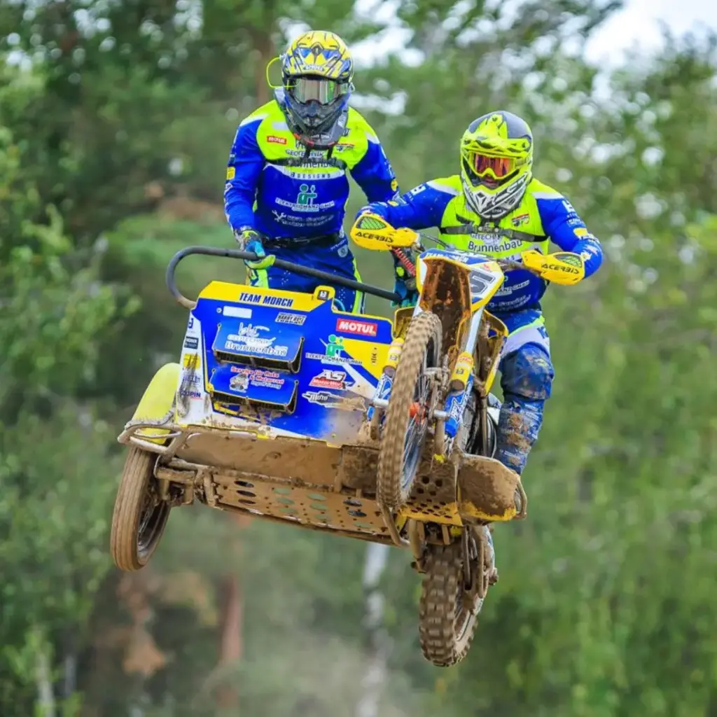 Motocross-Gespann mit Fahrer und Beifahrer Simon Hildebrandt (MSC Geisleden) springt in blau-gelber Rennmontur über eine schlammige Strecke.