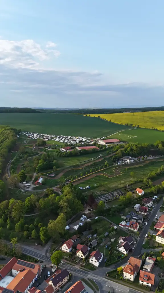 Luftaufnahme der Motocross-Anlage des MSC Geisleden mit Rennstrecke und belebtem Campingbereich, umgeben von Wald, grünen Ackerflächen und einem gelb blühenden Rapsfeld, neben einem Wohngebiet.