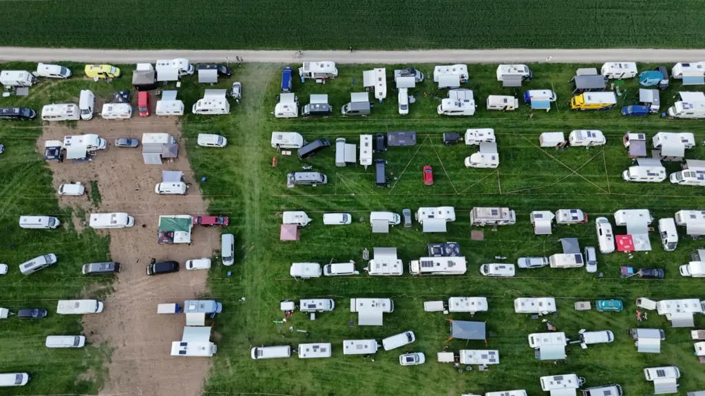 Luftaufnahme des weitläufigen Zelt- und Stellplatzes von MSC Geisleden, auf dem zahlreiche Wohnwagen, Wohnmobile und einige Zelte auf einem gemischten Untergrund aus Gras und Erde dicht beieinander stehen. Im Hintergrund ist ein grünes Feld und eine unbefestigte Straße zu sehen.