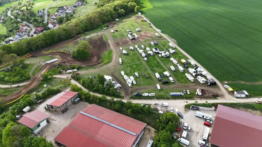 Luftaufnahme des Motocross-Geländes des MSC Geisleden mit einer kurvigen Erdpiste, einem großen Fahrerlager mit zahlreichen Wohnmobilen und Wohnwagen, umgeben von grünen Feldern, Waldstücken und einem Dorf im Hintergrund.
