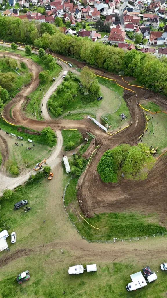 Luftaufnahme der MSC Geisleden Motocross-Strecke mit gewundenen Erdpisten, umgeben von grünen Wiesen und Bäumen. Im Hintergrund ist ein Dorf mit Häusern und roten Dächern zu sehen. Auf dem weitläufigen Gelände sind mehrere Fahrzeuge, temporäre Strukturen und Personen verteilt, was auf eine Veranstaltung hinweist.